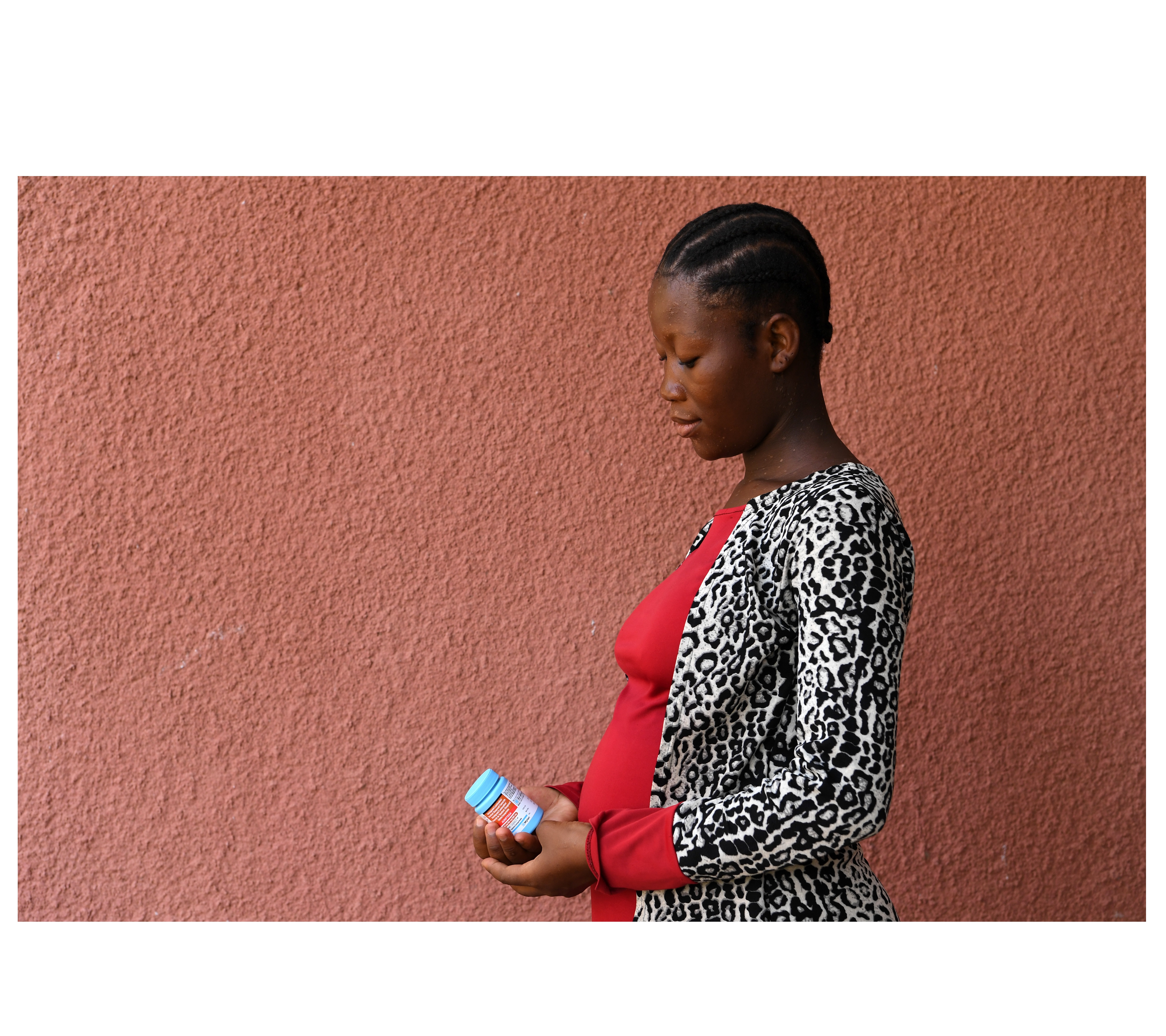 A 20 year-old pregnant woman living with HIV is standing against a textured pink wall, looking down thoughtfully at a bottle of medication in her hands