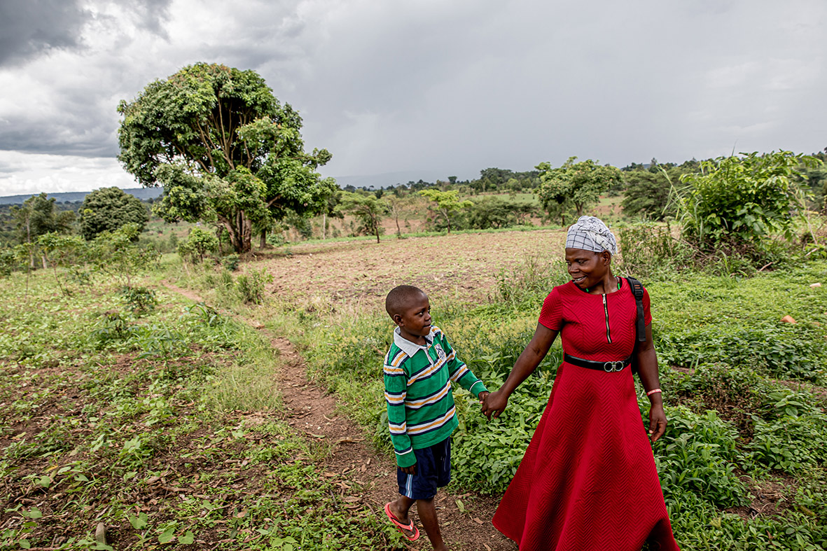 This image shows a rural scene with a woman and a child walking through a field. The two are holding hands as they walk along a dirt path surrounded by greenery.