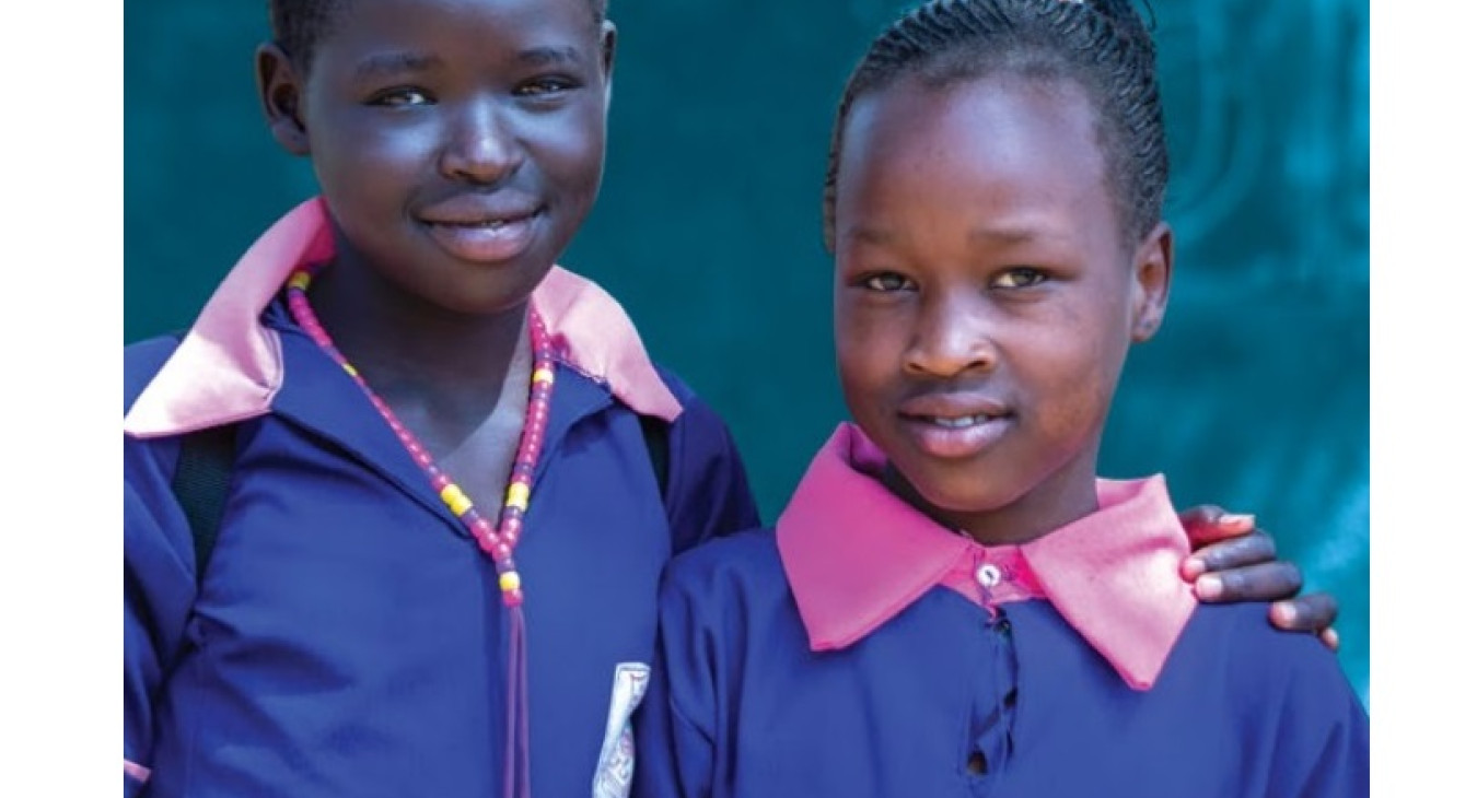 The photo features two young girls. One is carrying a backpack and has her hand over the shoulder of the other, who is holding a notebook.