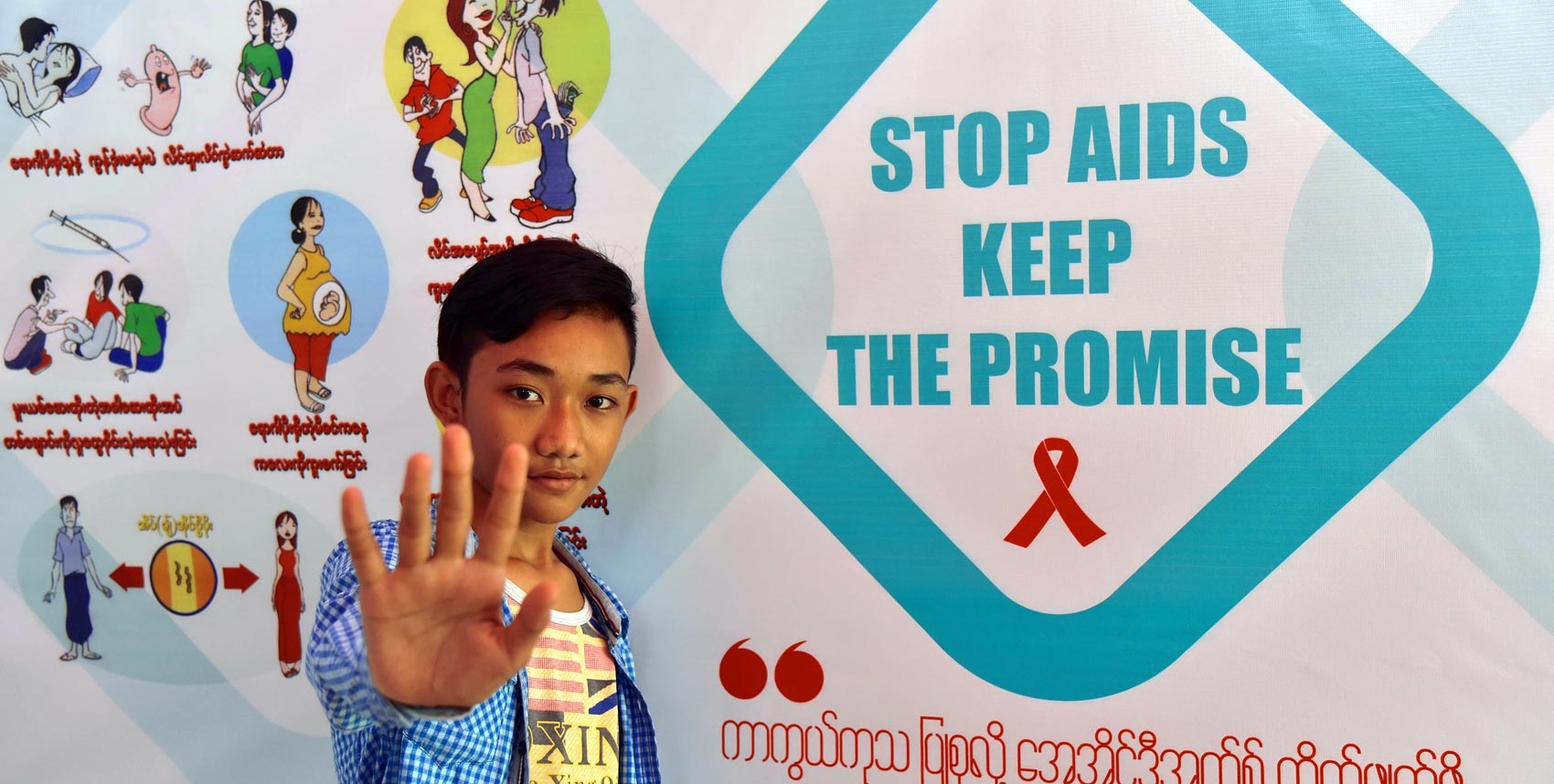 A young man gestures stop in front of a poster that says ‘Stop AIDS Keep the Promise’